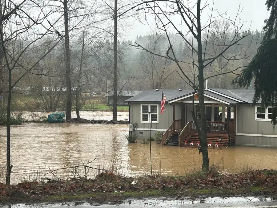 A home surrounded by brown creek flood water under a rainy wooded hillside, creek flooding near Gresham, OR